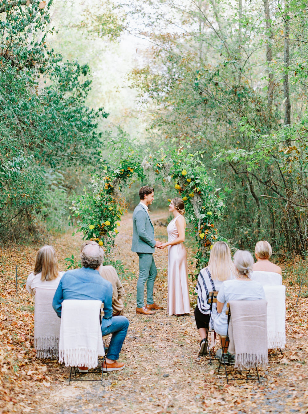 autumn wedding ceremony with a foraged flower arch, groom in sage green suit and bride in apricot silk gown