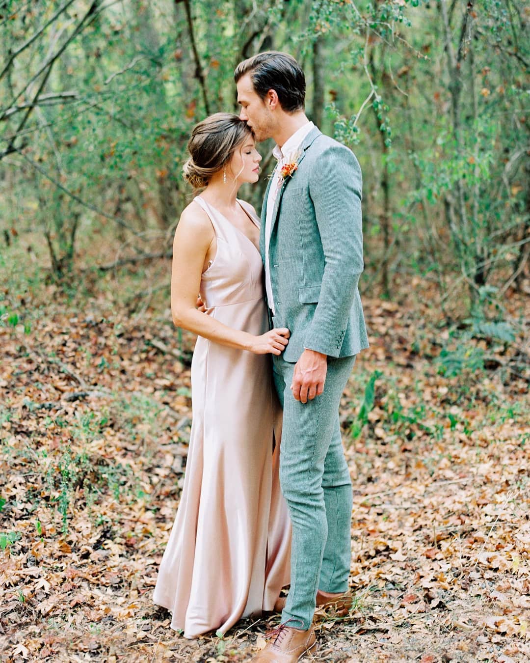 Bride in a silk apricot gown and groom in a sage green wool suit sharing a forehead kiss in an autumn forest in Columbia, Charleston SC