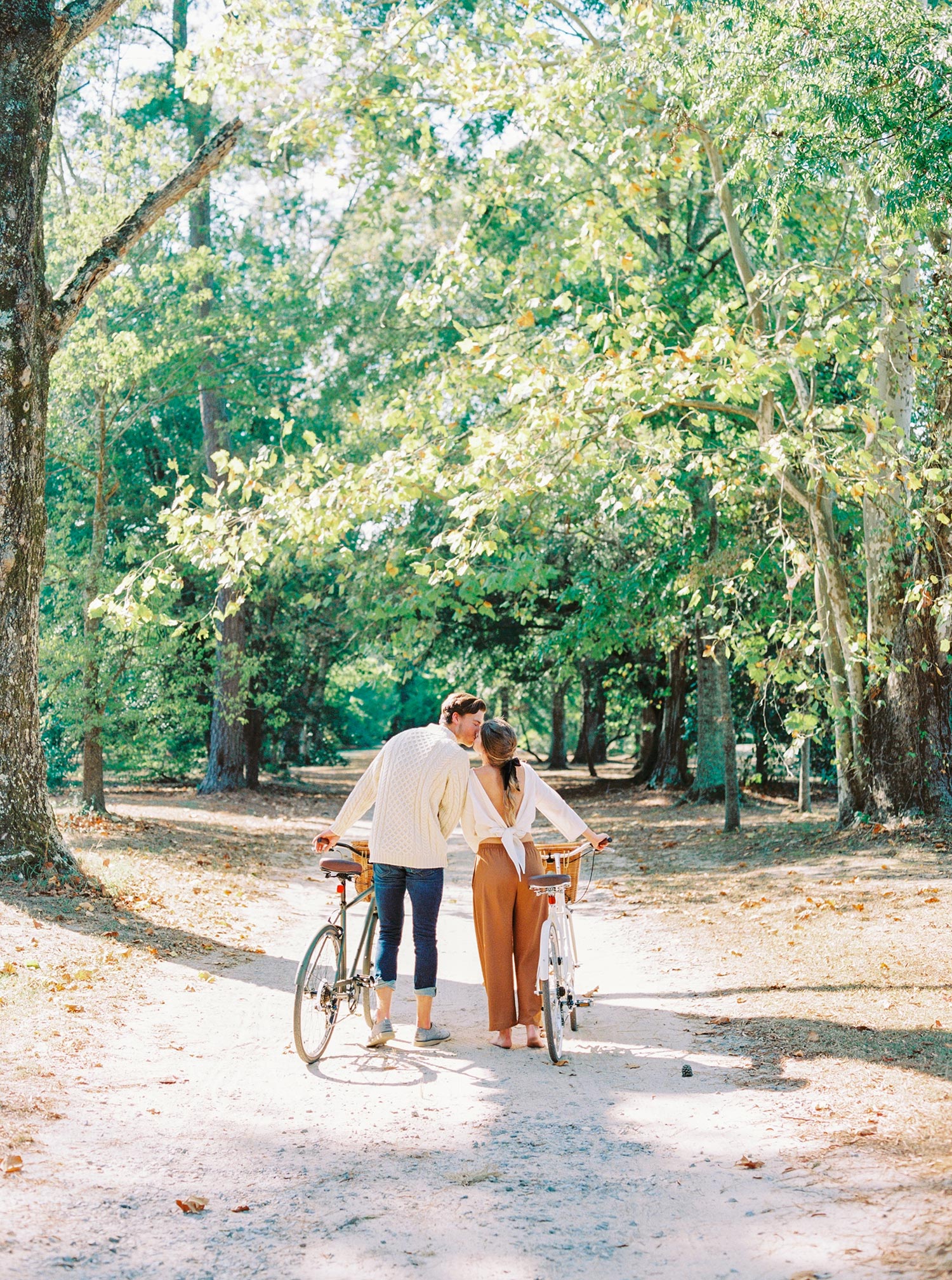 white linen blouse with rust colored pants and a fisherman sweater walking their bicycles down an open autumn forest path