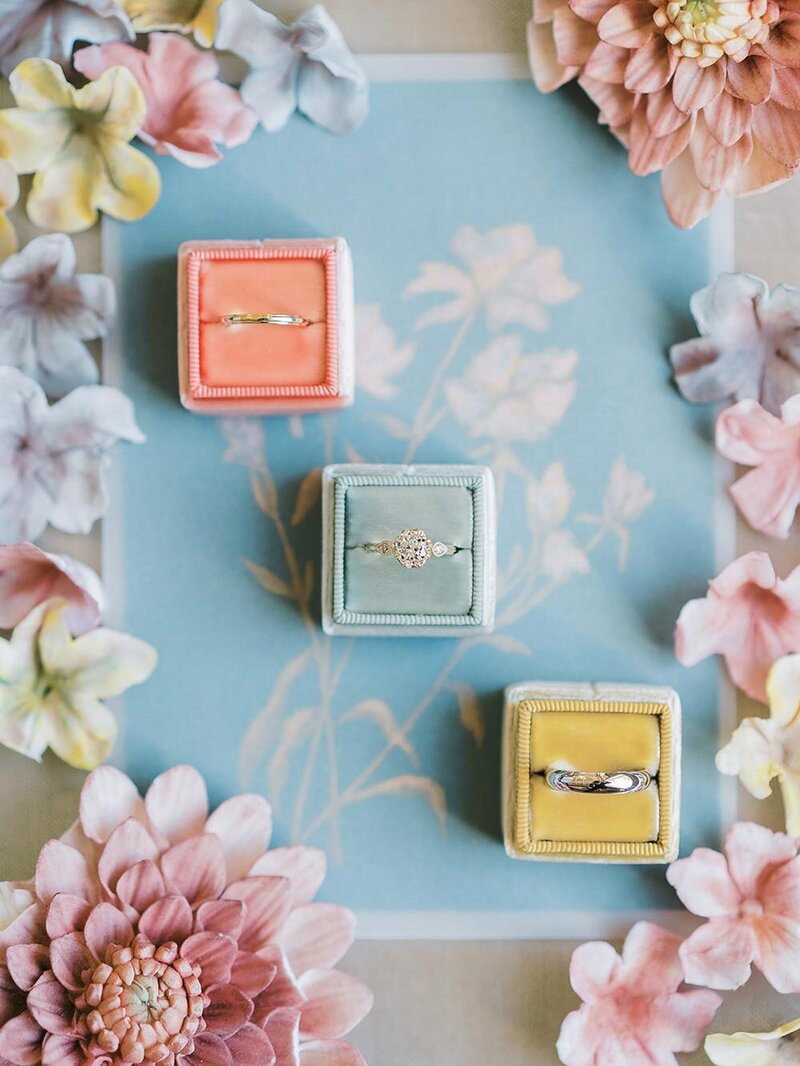 peach, sage and mustard velvet ring boxes with a halo engagement ring surrounded by pastel sugar flowers at a Charleston wedding, with flatlay styling by Willow and Oak Events and photography by Kylee Yee