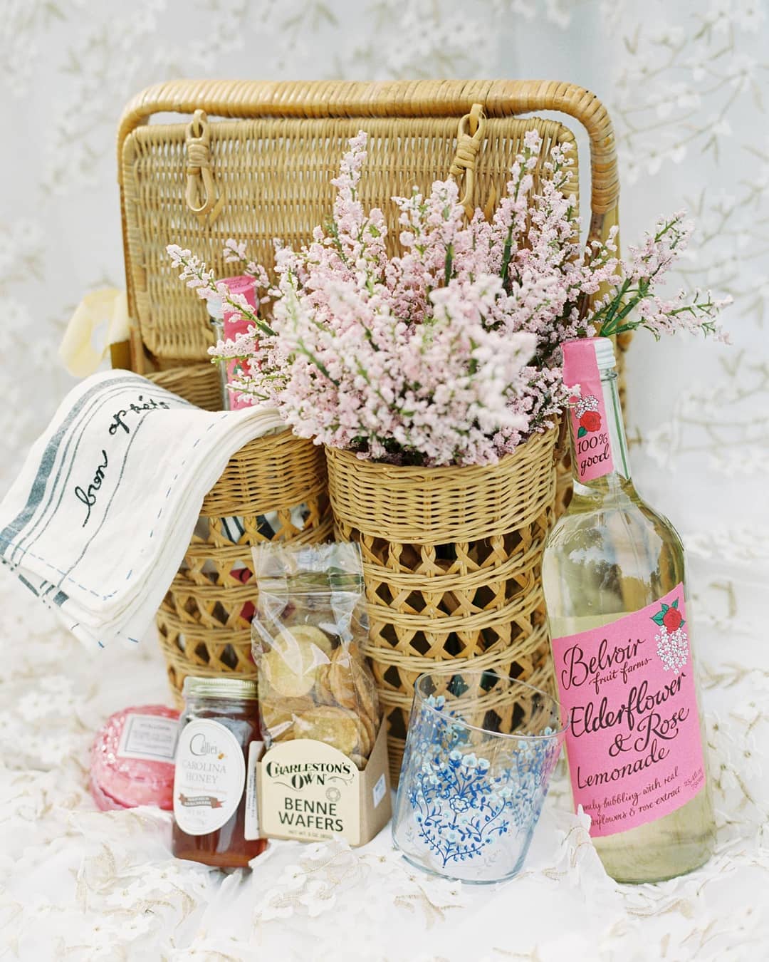 rattan picnic basket with wine bottle holders for unique wedding welcome gift packaging, holding flowers, benne wafers, honey, Belvoir elderflower, linen napkins, handpainted glassware and a peony candle for a destination wedding in Charleston, SC at Middleton Place; photography by The Happy Bloom and planning by Willow and Oak Events