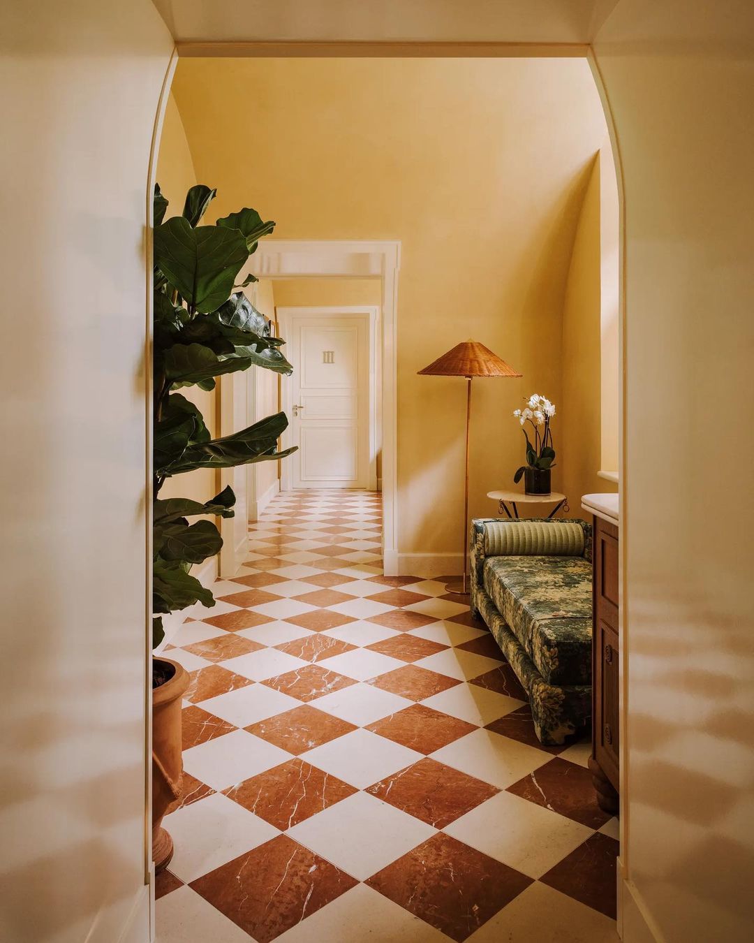 A brown and white checkered floor in the corridor leading to the Guerlain Spa at Saint James Paris, accented by a green pastoral patterned chaise lounge and skinny rattan floor lamp