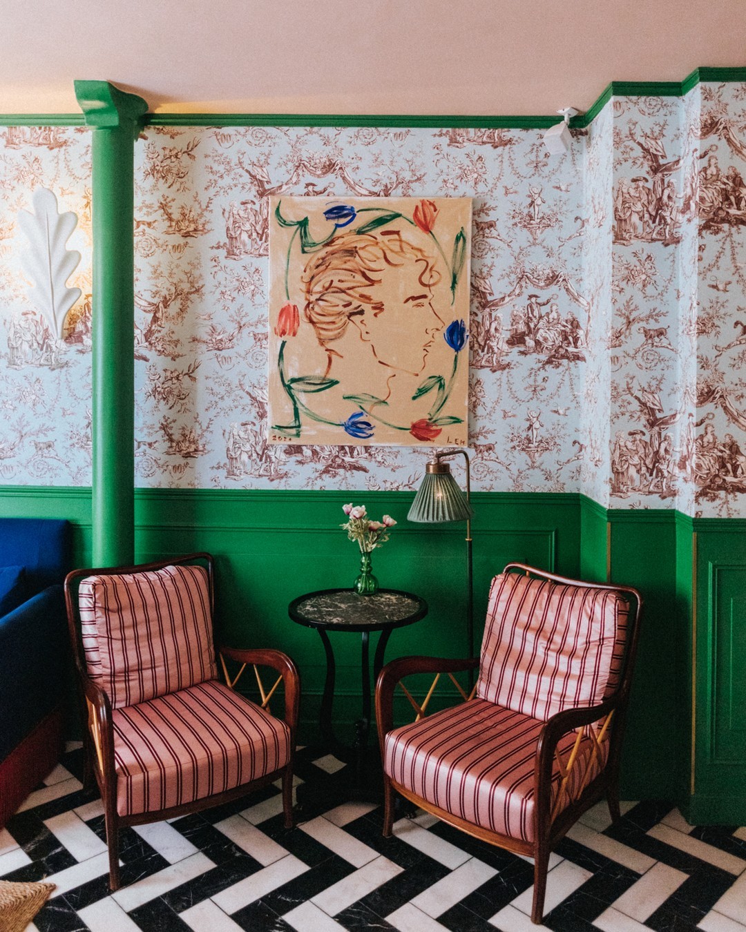 The eclectic mixed print lobby design of Hotel Les Deux Gares in Paris by Luke Edward Hall with striped pink + red satin armchairs, black + white marble zig zag floors, a blue + brown toile de jouy fabric wall split by glossy green paneled wainscoting and a Greco-Roman inspired bust portrait from the artist himself