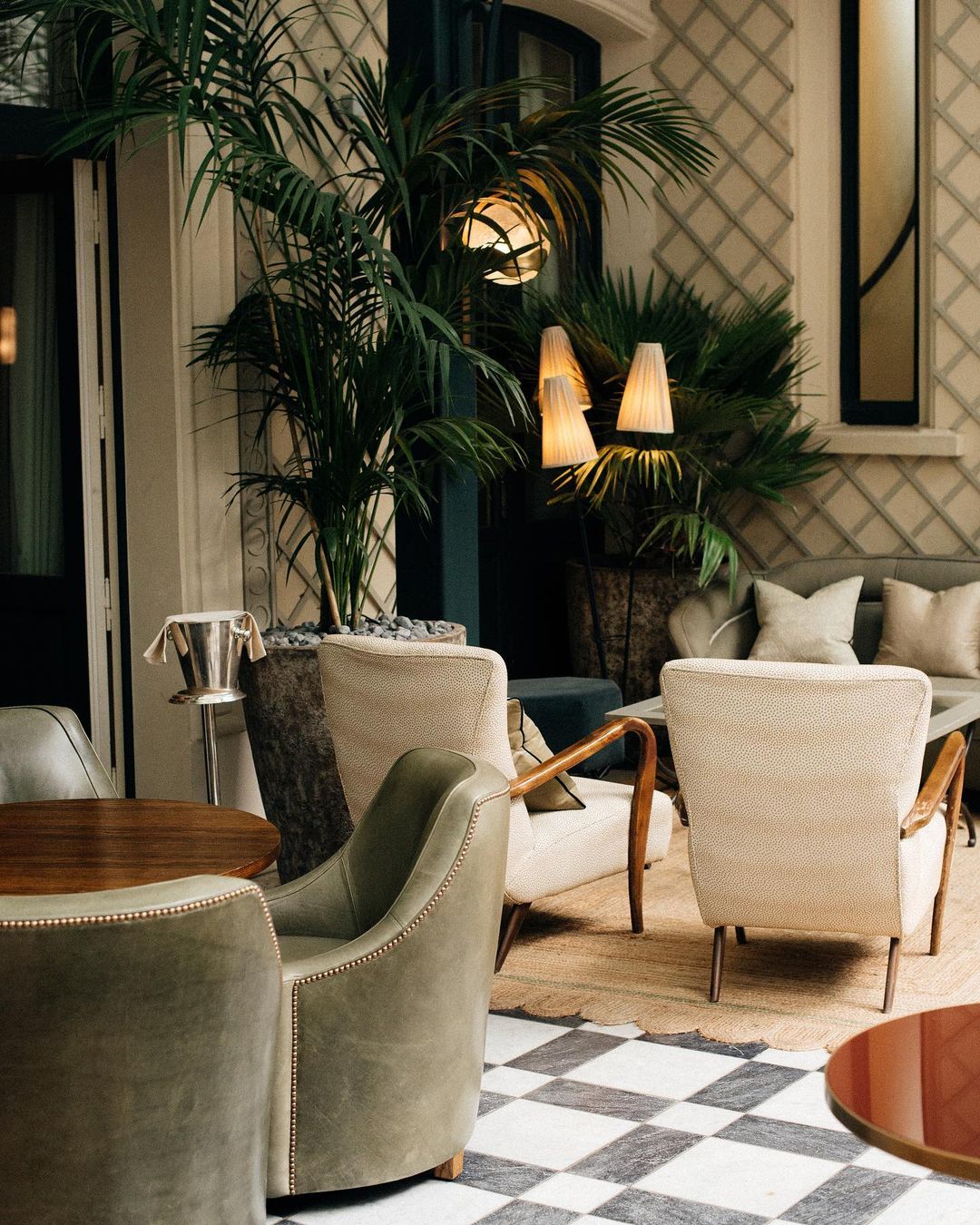 A sage and cream colored lounge space at Soho House Paris with brushed leather chairs, black and white checkered floors, scalloped jute rugs, latticework walls and floor-to-ceiling plants