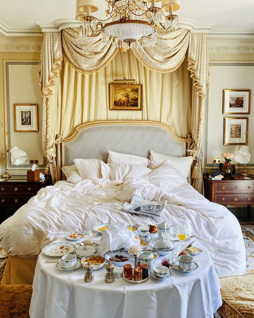 An indulgent breakfast-in-bed scene at Ritz Paris with a gold canopy, ticking stripe headboard, antique wooden furniture and framed gallery art