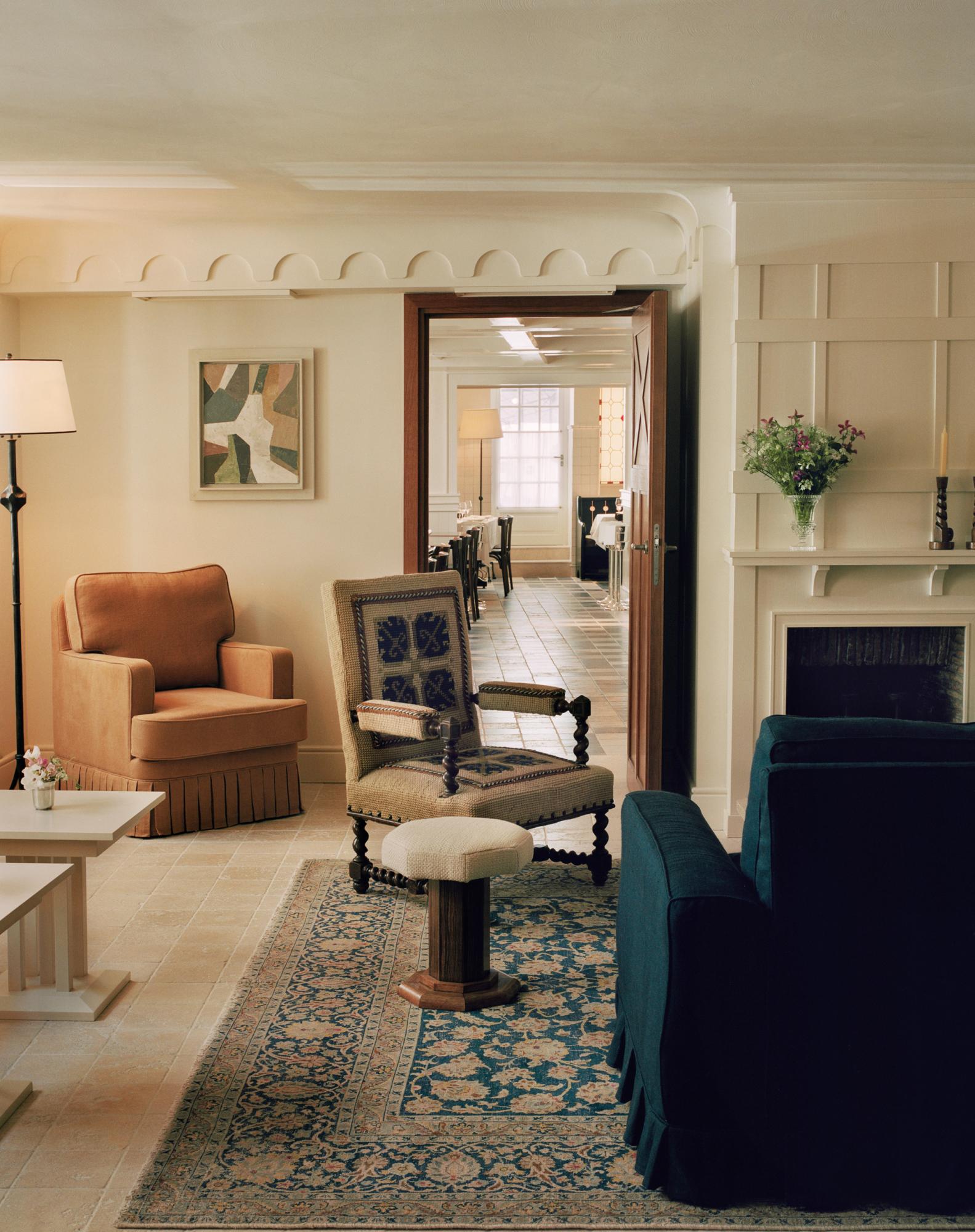 The lounge of Chateau Voltaire with a vintage Italian needlework side chair, a fringed leather armchair and a ruffled skirt sofa in a deep peacock blue