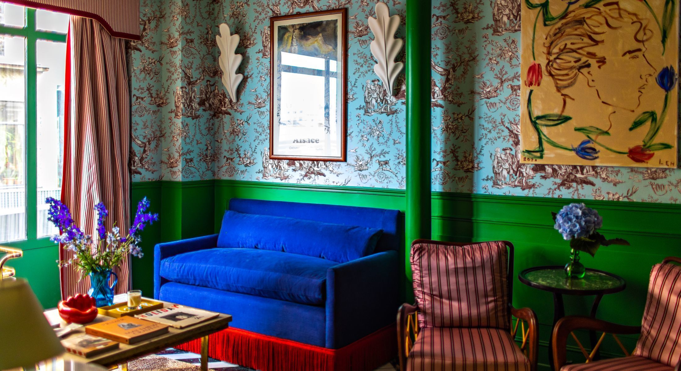The eclectic mixed print lobby design of Hotel Les Deux Gares in Paris by Luke Edward Hall with striped pink + red satin armchairs, an electric blue velvet sofa with bright red fringe, leafy wall sconces inspired by Matisse, a blue + brown toile de jouy fabric wall split by glossy green paneled wainscoting and a Greco-Roman inspired bust portrait from the artist himself