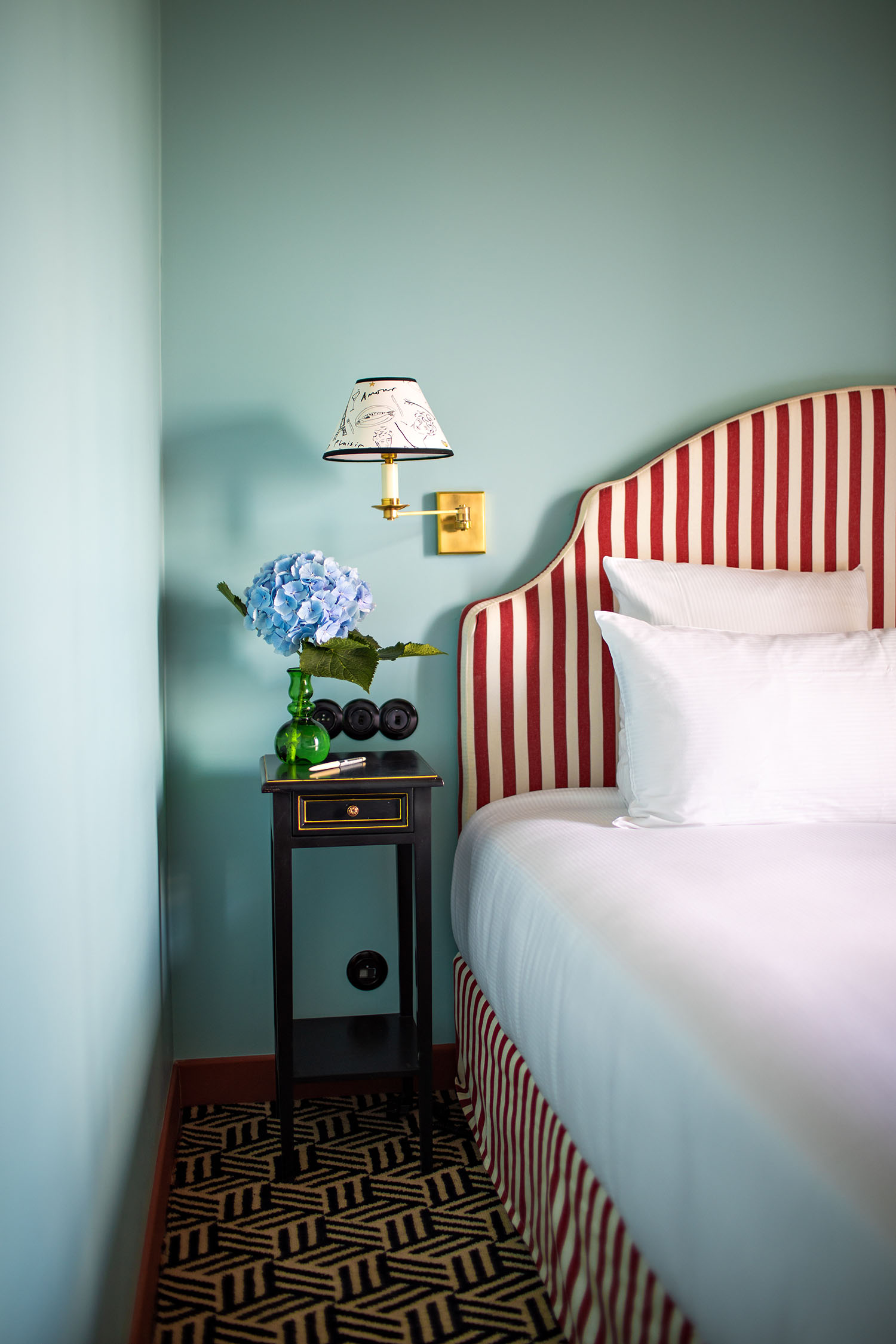 An ice blue guest room at Hotel les Deux Gares with a red + white striped headboard, geometric patterned carpeting, amural-sketched wall lamp shades, and a small column-shaped side table in a glossy mahogany wood