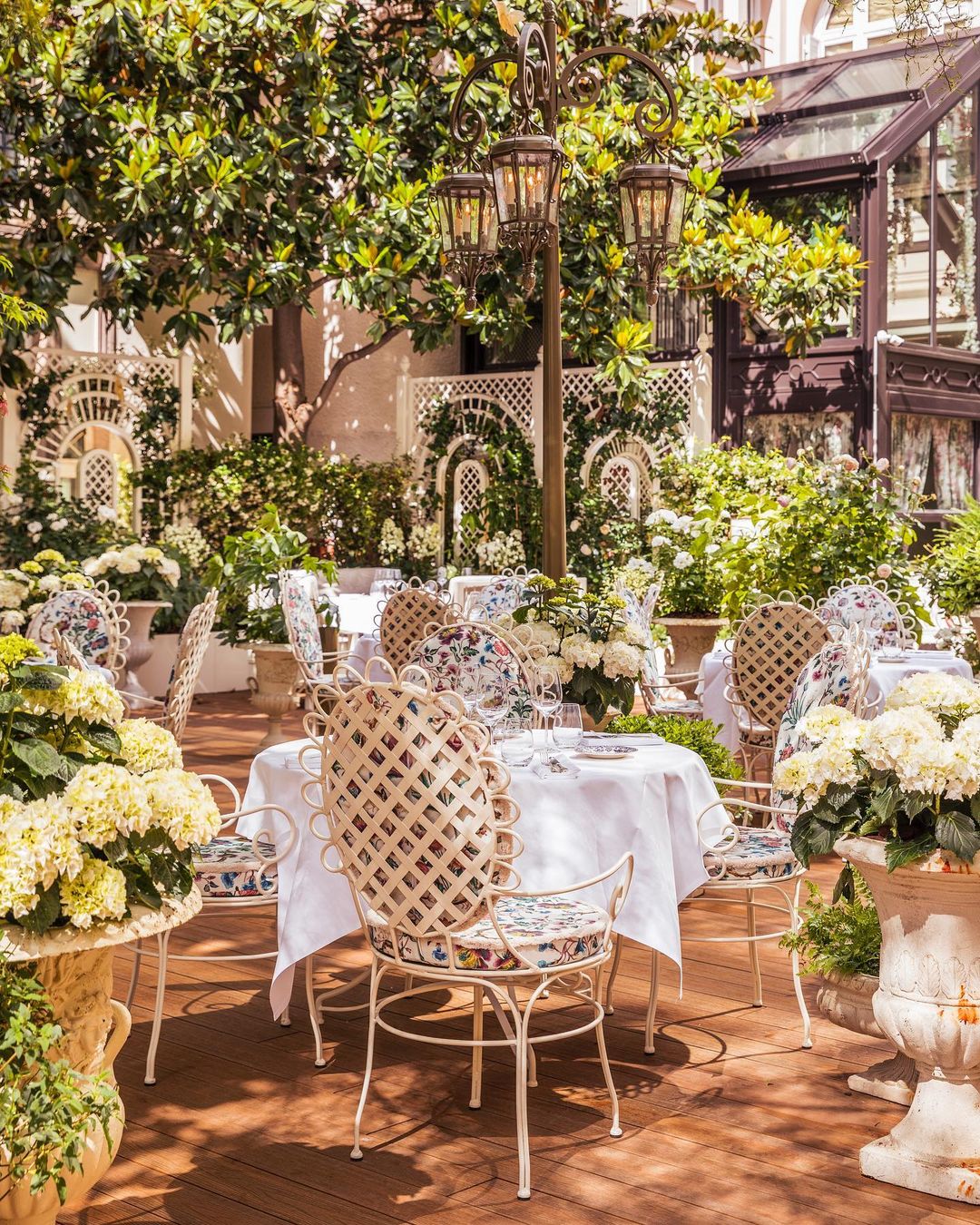 Maison Russe garden terrace dining with white iron lattice-backed chairs featuring a scalloped iron trim and custom floral fabric wrapped around the cushions