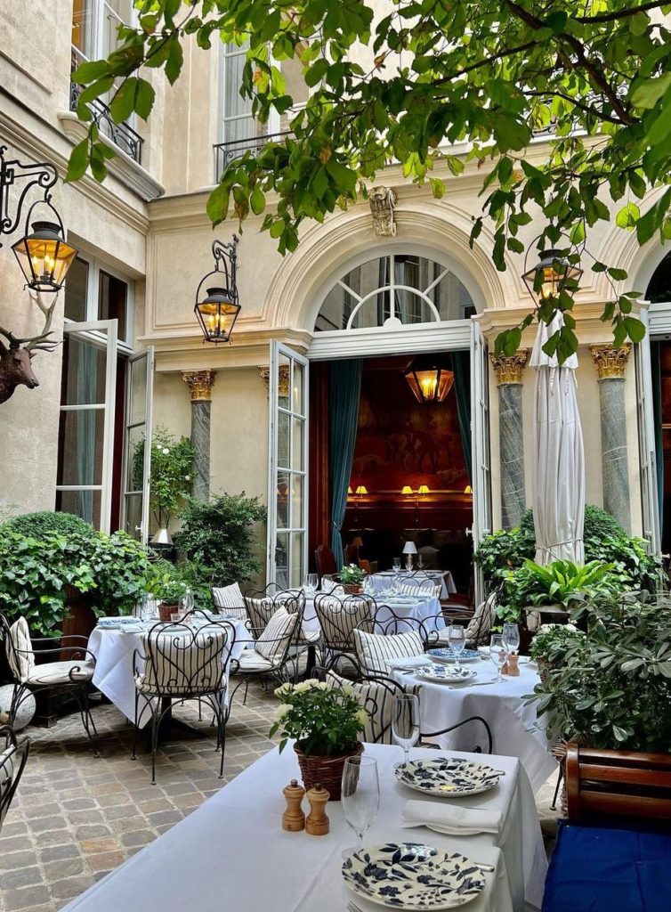 The secret garden aesthetic of Ralph's Paris with black iron garden chairs, striped blue + white pillow cushions, potted plant centerpieces, and mismatched floral plates