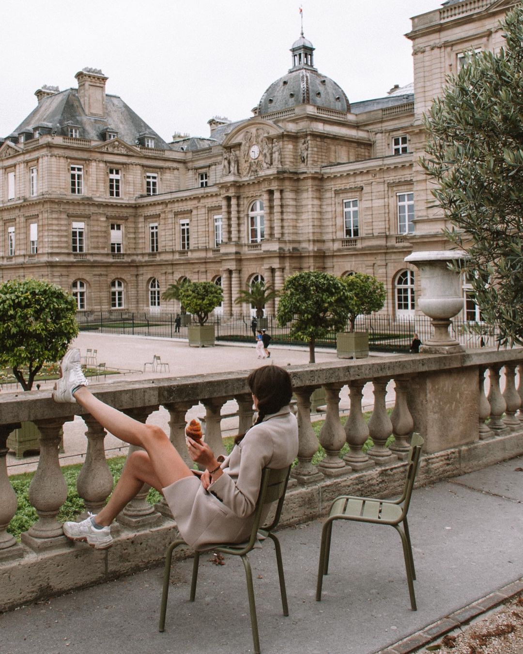 A woman lounges with a croissant in hand on one of the sage metal garden chairs at Jardin du Luxembourg