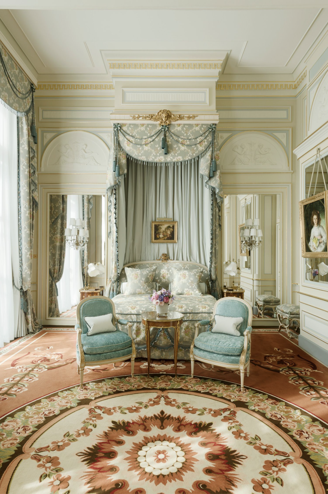 The blue and cream Suite Imperiale at Ritz Paris with a lavish canopy bed, floral print bedding, velvet teal arm chairs, mahogany tables and a large coral and green rug that extends the length of the room
